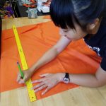 Picture of a student working on a project in the Innovation and Collaboration Studio, using a yardstick to draw a straight line on orange material