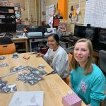 Picture of two students sitting at a table and working on a project in the Innovation and Collaboration Studio; table covered with various tools and materials