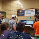 Picture of a group of students with their backs to the camera, gathered around and looking at a computer screen in the Innovation and Collaboration Studio
